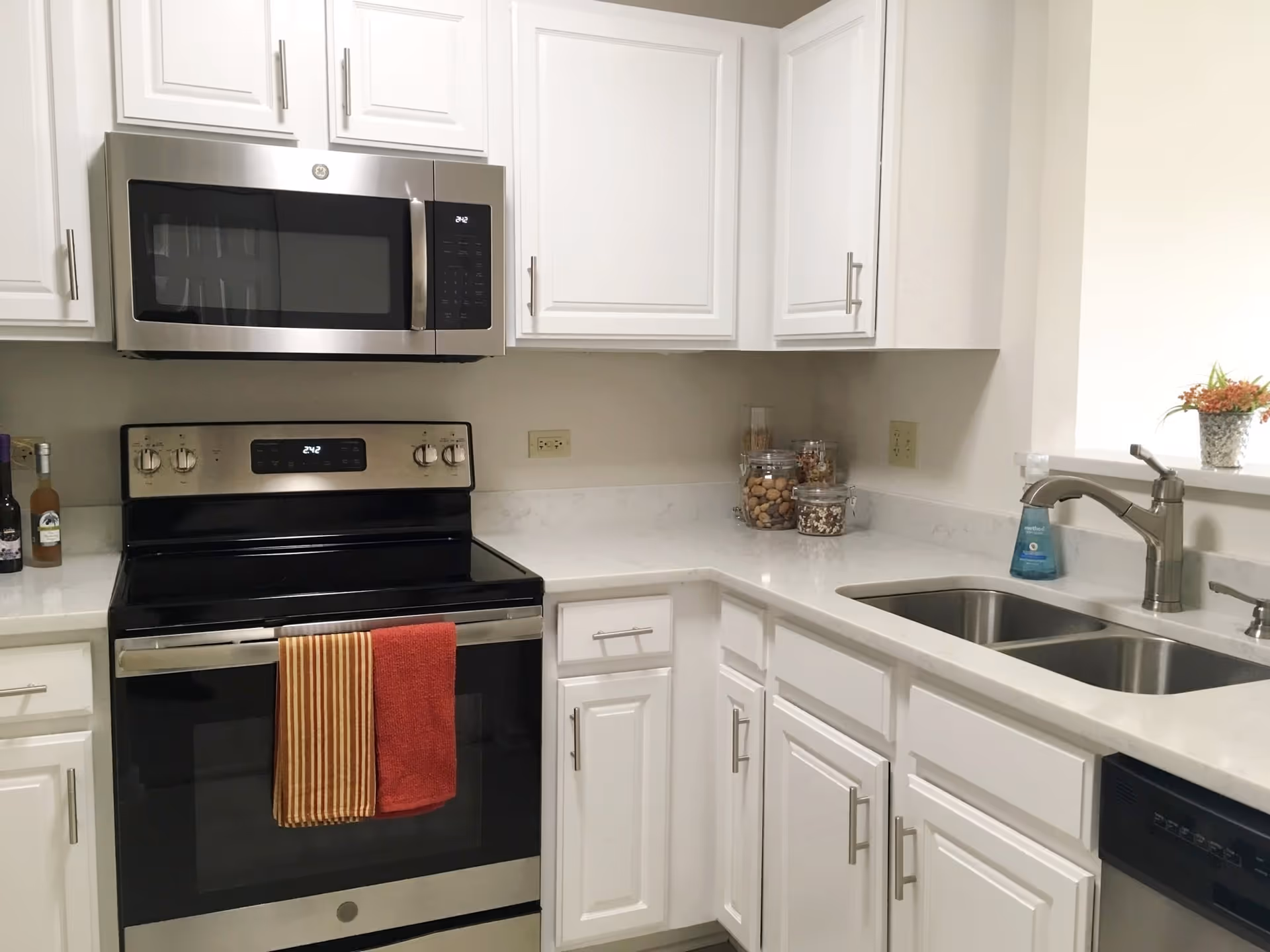 A modern kitchen with white cabinets, a stainless steel microwave and oven, a double sink with a faucet, and a countertop with jars containing nuts and snacks. Two towels hang on the oven handle, and a small plant is placed on the counter near the sink.