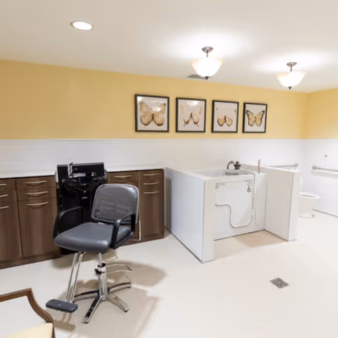 A spacious senior living facility room with a salon chair and hair washing station on the left, a walk-in bathtub with safety rails in the center, and a toilet area partially visible on the right. The walls are painted yellow and white, with four framed butterfly pictures hanging above the bathtub. The floor is tiled in light beige, and the room is well-lit with ceiling lights.