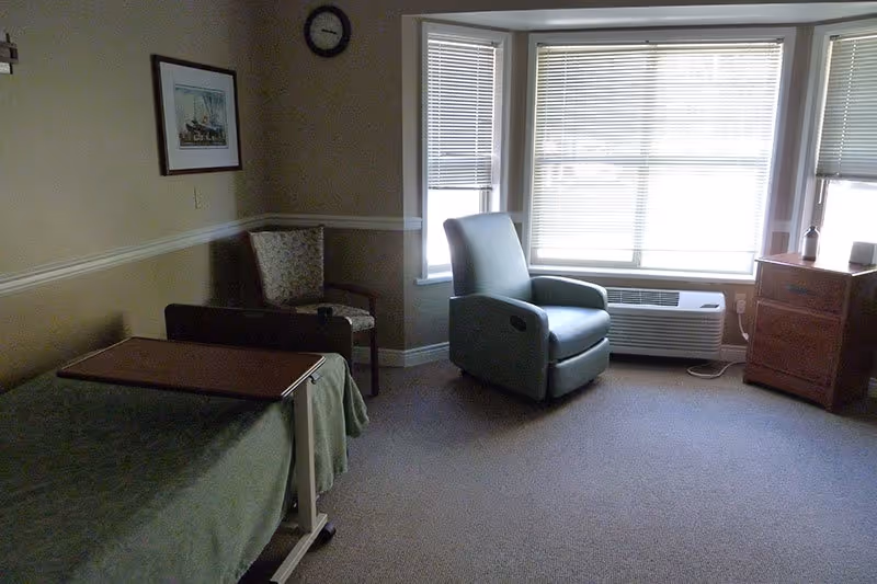 A room with a single bed covered with a green blanket and an overbed table, a patterned armchair in the corner, a recliner chair near a large window with blinds, a wooden nightstand with items on top, beige walls with a framed picture and a clock, and carpeted floor.
