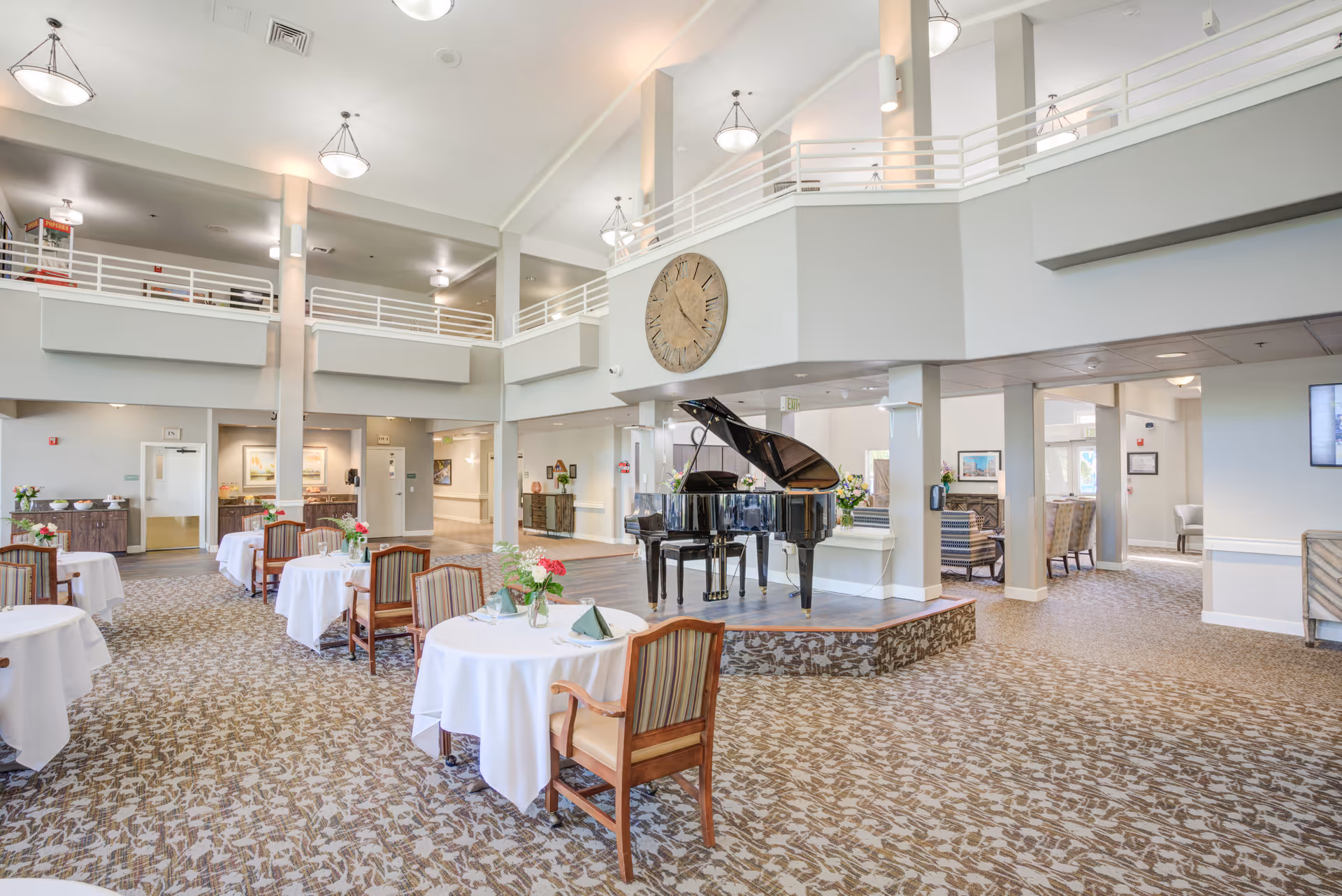 Spacious dining area in a senior living facility with round tables covered in white tablecloths, each set with flowers and napkins. A black grand piano is placed on a raised platform in the center. The room has high ceilings with modern light fixtures and a large clock on the wall above the piano. There is a second-floor balcony overlooking the dining area and comfortable seating areas visible in the background.