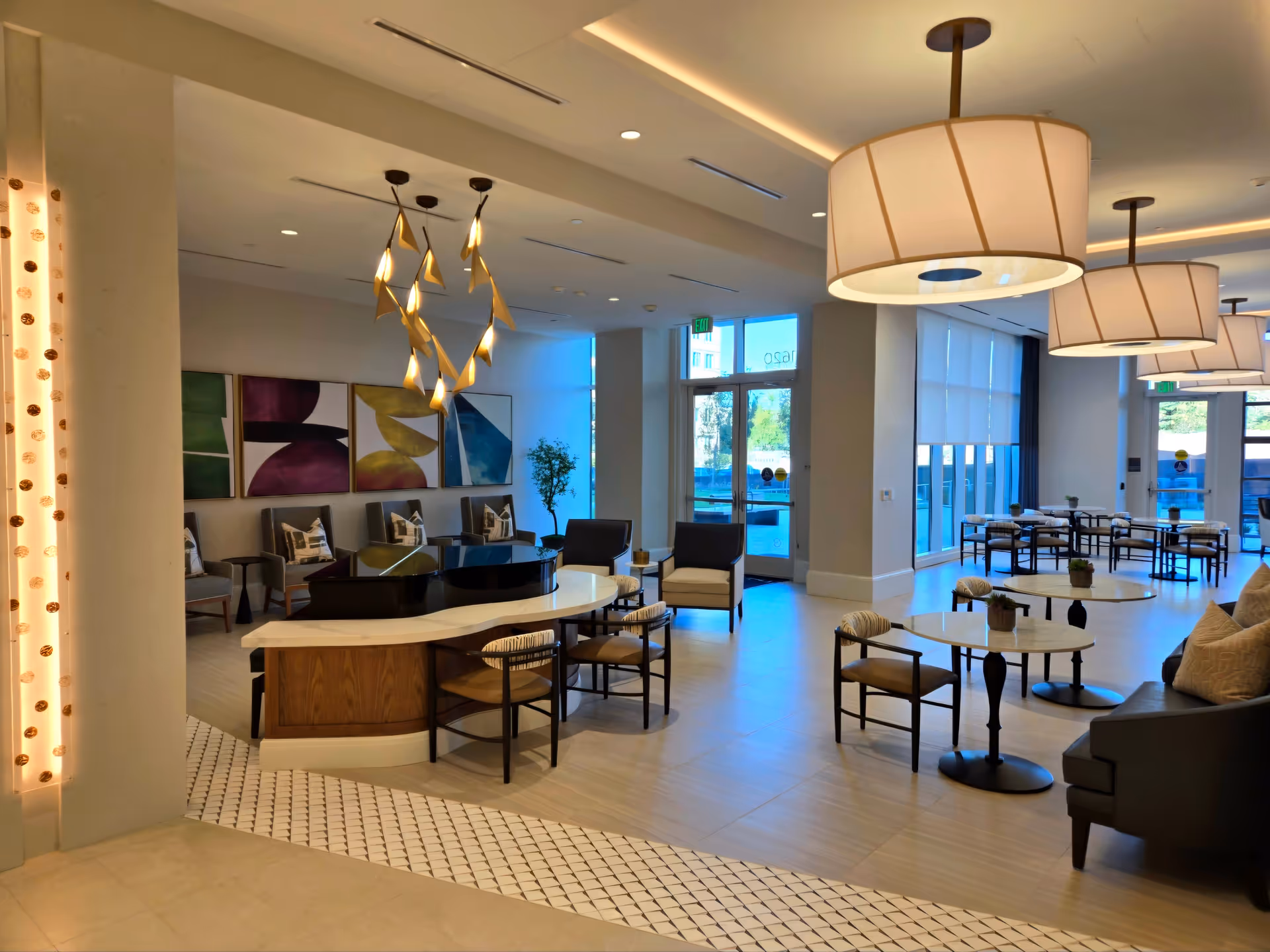 A spacious and modern common area in The Trillium facility featuring a black grand piano with a white marble top, surrounded by chairs. The room has large windows allowing natural light, contemporary pendant lights hanging from the ceiling, abstract artwork on the wall, and several small tables with chairs arranged for seating.