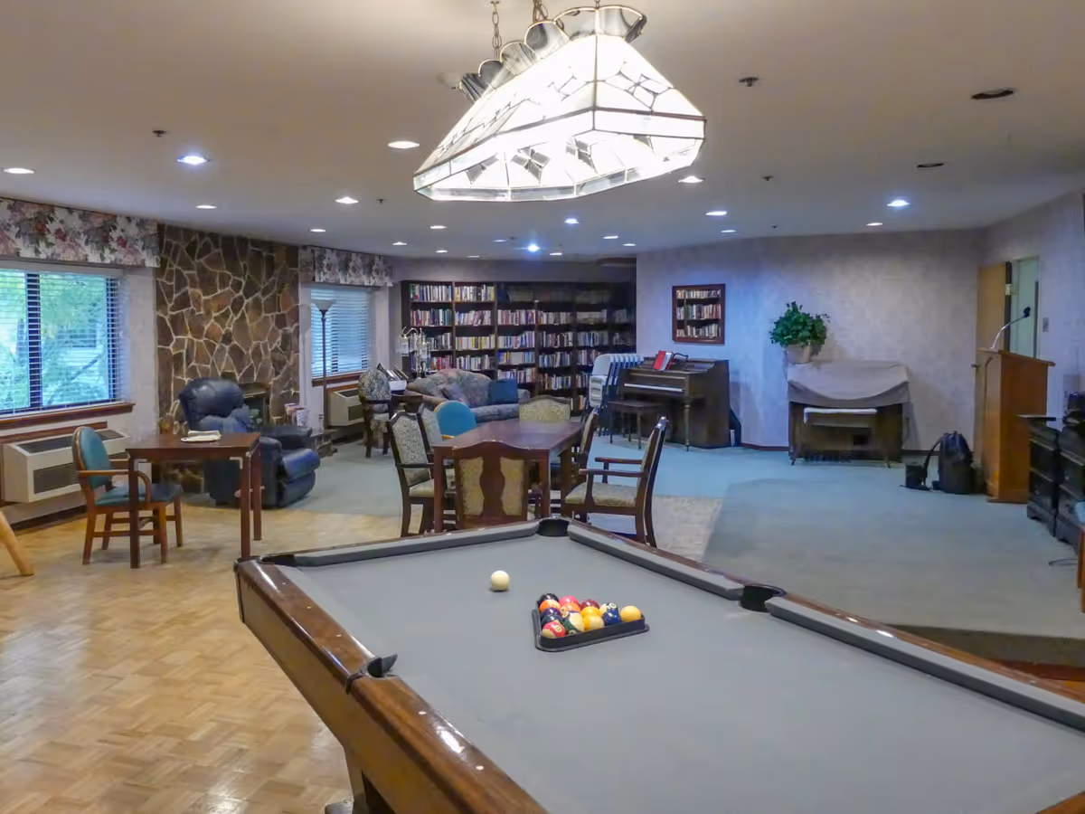 A spacious senior living common area featuring a pool table in the foreground with billiard balls arranged for a game. The room has a wooden floor section and carpeted area with several chairs and tables. There is a stone fireplace on the left side, large windows with floral valances, bookshelves filled with books, a piano, and a covered organ. The ceiling has recessed lighting and a large decorative hanging light fixture.