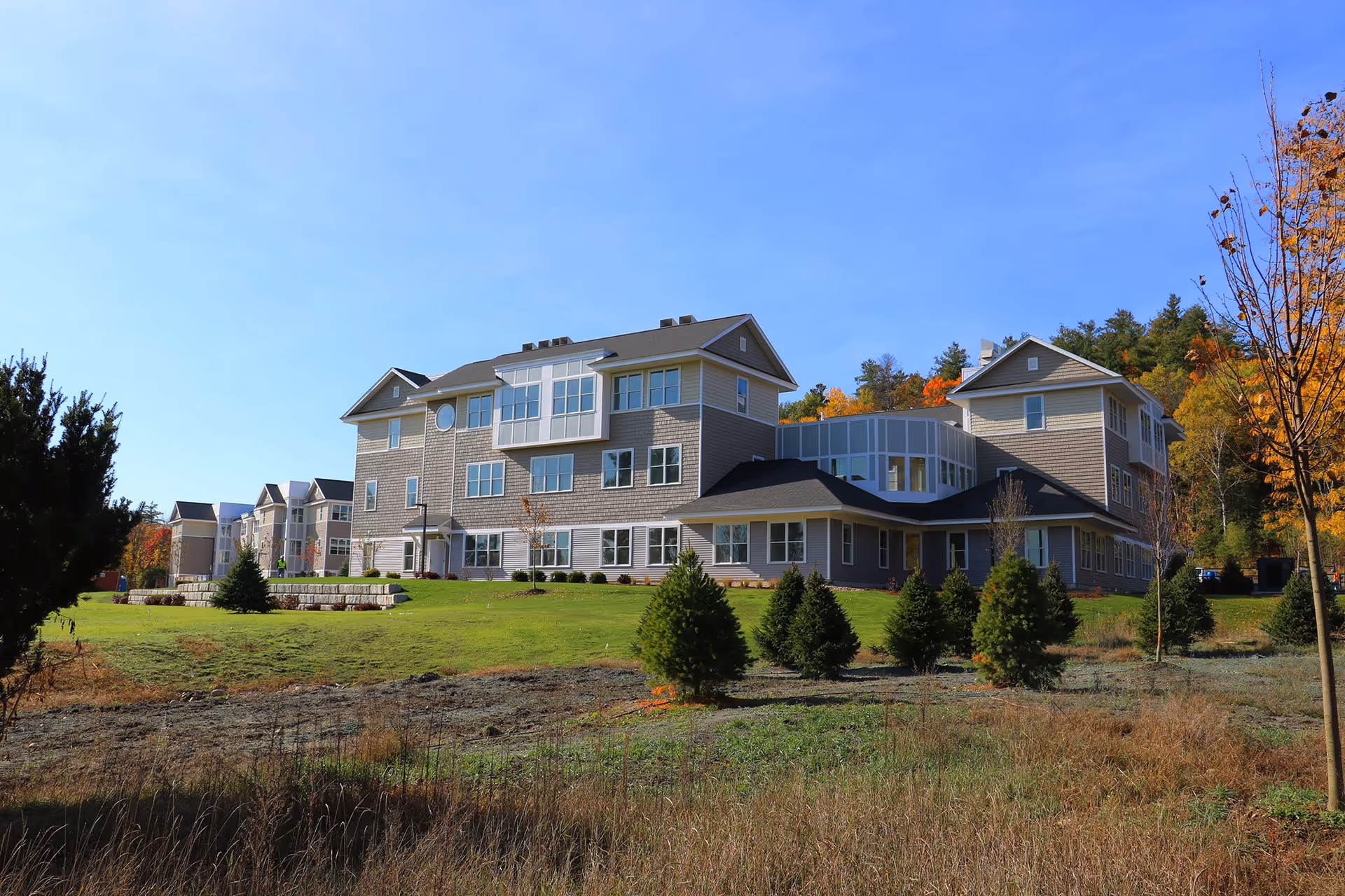 Exterior view of a large, multi-story senior living facility building with gray siding and numerous windows, surrounded by a well-maintained lawn, small evergreen trees, and autumn foliage under a clear blue sky.