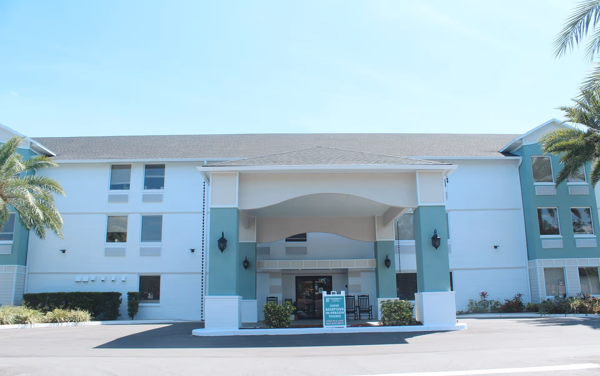 Front exterior view of a three-story senior living facility building with a covered entrance supported by columns. There are palm trees on both sides and a sign near the entrance indicating that in-person tours are now being accepted.