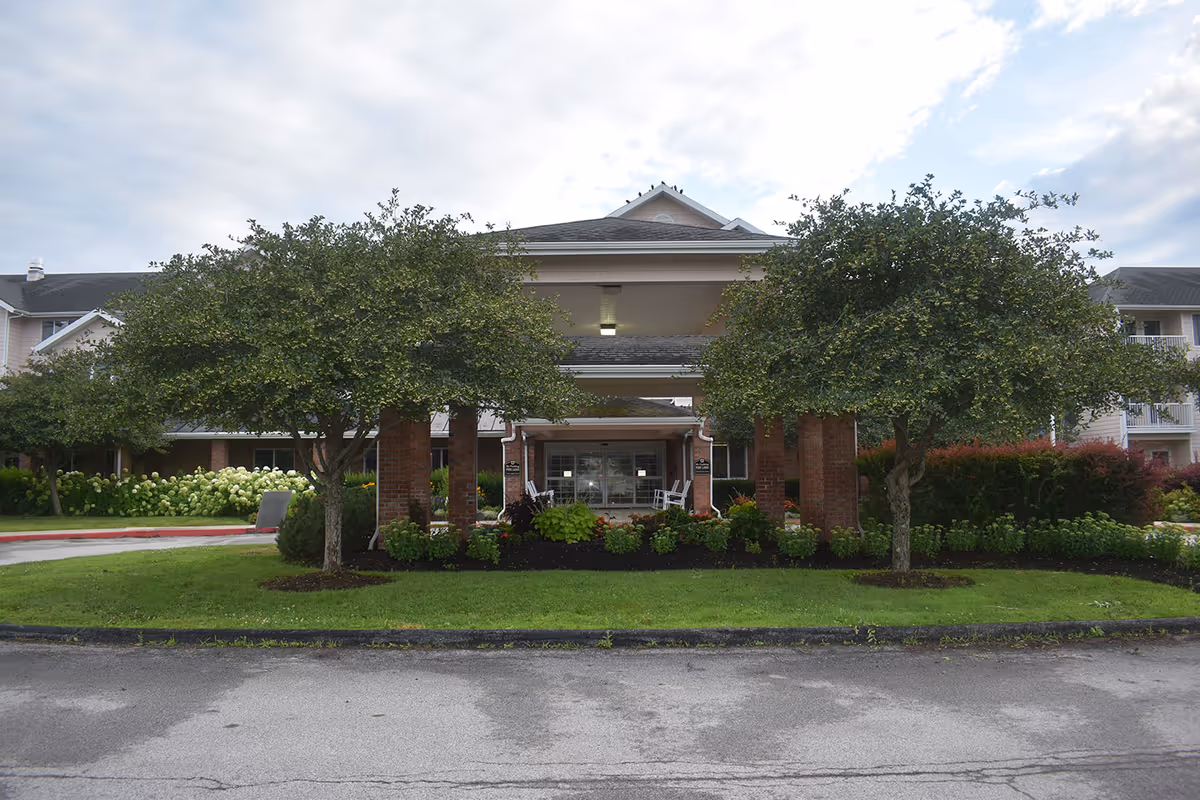 Front exterior view of Solstice Senior Living at Bangor showing the entrance with a covered driveway supported by brick pillars, flanked by two trees and landscaped greenery under a partly cloudy sky.
