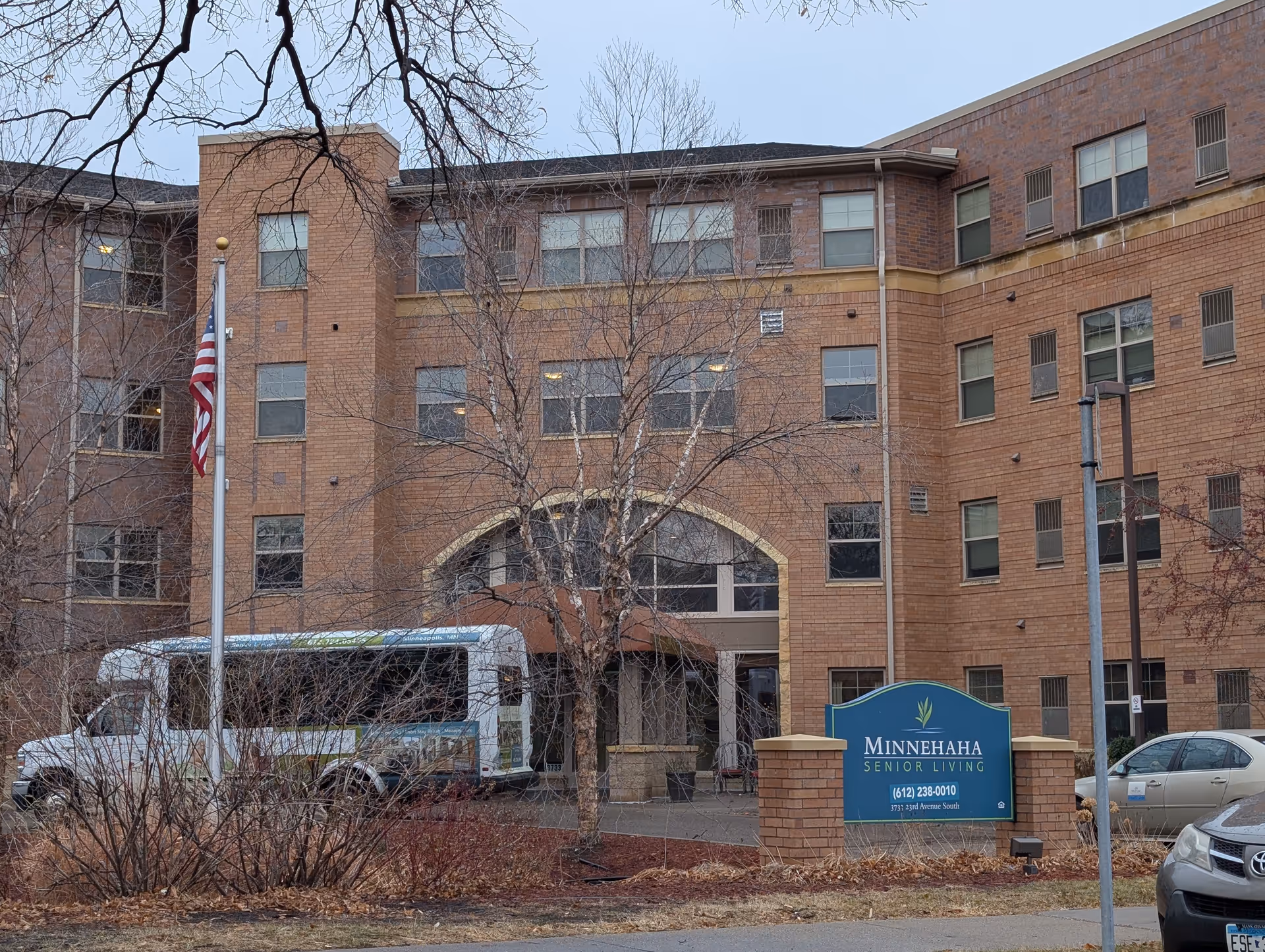Exterior view of Minnehaha Senior Living, a multi-story brick building with several windows. There is a blue sign in front displaying the facility's name and contact information. An American flag is on a flagpole near the entrance, and a white shuttle bus is parked in front of the building. Leafless trees and shrubs are visible in the foreground.