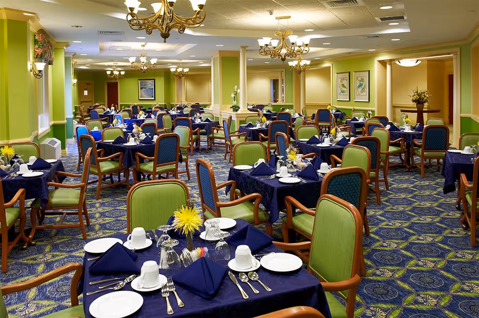 Spacious dining room with multiple round tables set with navy tablecloths, green upholstered chairs, chandeliers, and a patterned carpet.