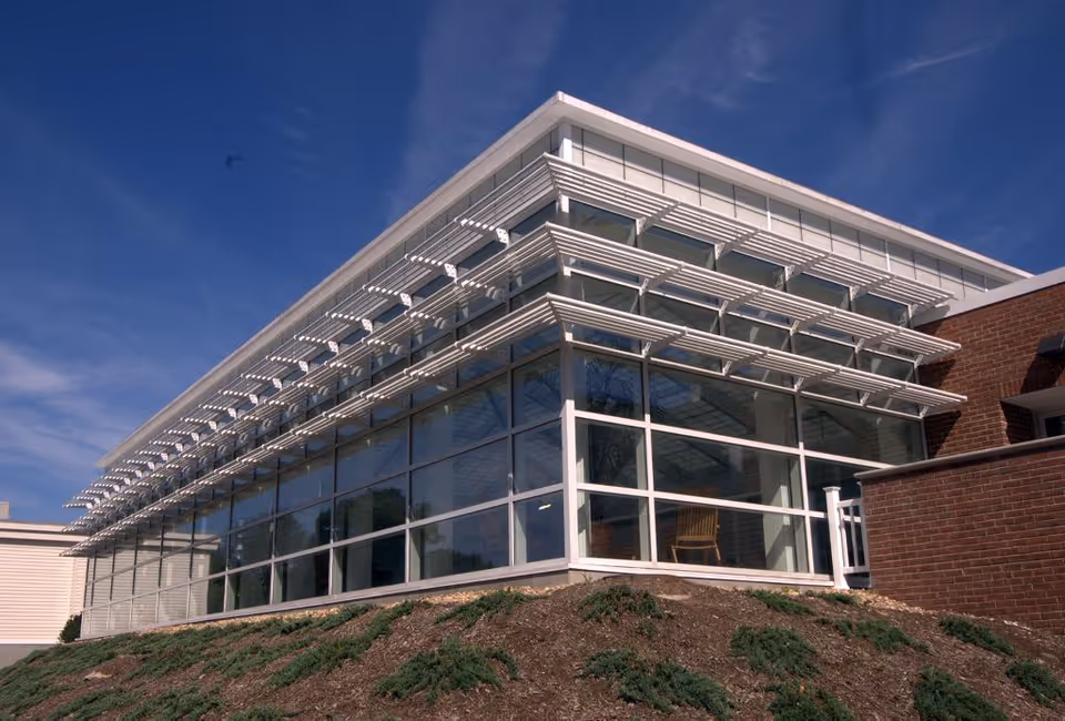 Corner view of a modern glass-front building with horizontal sunshades, brick accents, and a small landscaped mound under a blue sky.