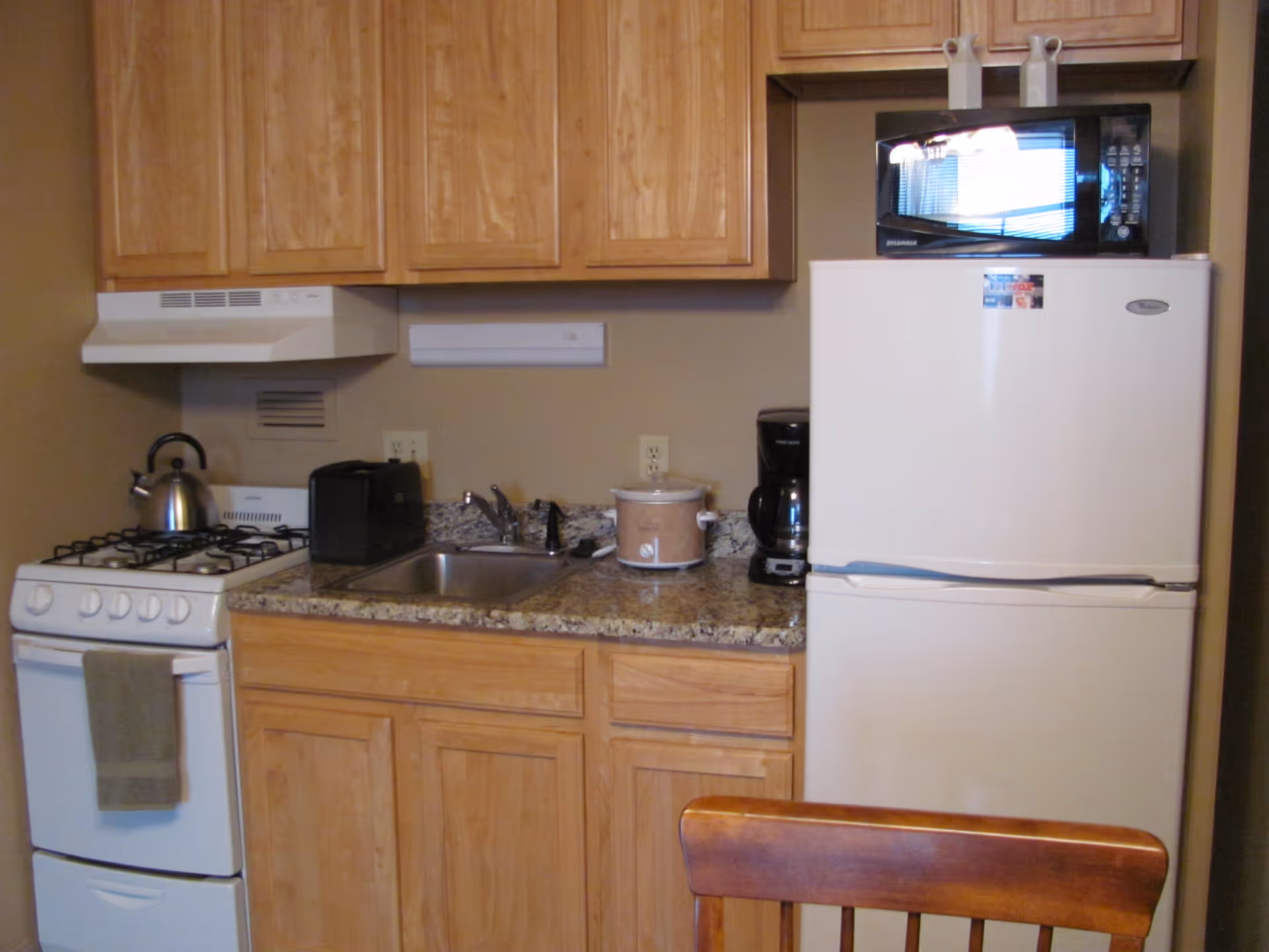 Small kitchen area with wooden cabinets, a white gas stove with a kettle on top, a granite countertop with a sink, a toaster, a slow cooker, a coffee maker, a white refrigerator with a microwave on top, and a wooden chair partially visible in the foreground.
