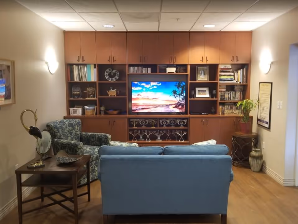 A cozy seating area with a blue sofa and patterned armchair facing a wooden built-in entertainment center with a TV and bookshelves.