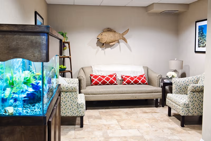 A cozy sitting area with a beige sofa adorned with two red patterned pillows, two patterned armchairs, a wooden side table with a lamp and flowers, a large fish tank on the left, and a decorative woven fish mounted on the wall.
