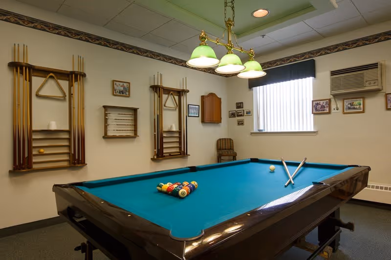 Interior room with a pool table set up for a game, two pool cues resting on the table, and a rack of pool cues mounted on the wall. The room has beige walls with framed pictures, a window with vertical blinds, an air conditioning unit, and a ceiling light fixture with four green lampshades.