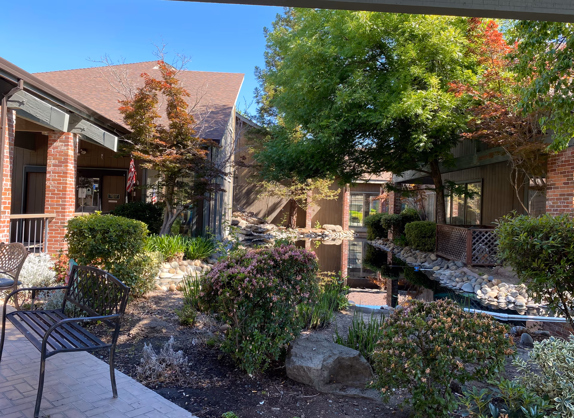 Outdoor courtyard area at The Crest at Citrus Heights featuring a small pond with rocks along the edges, surrounded by bushes, trees with green and red leaves, and brick buildings. There is a metal bench on a paved walkway in the foreground.