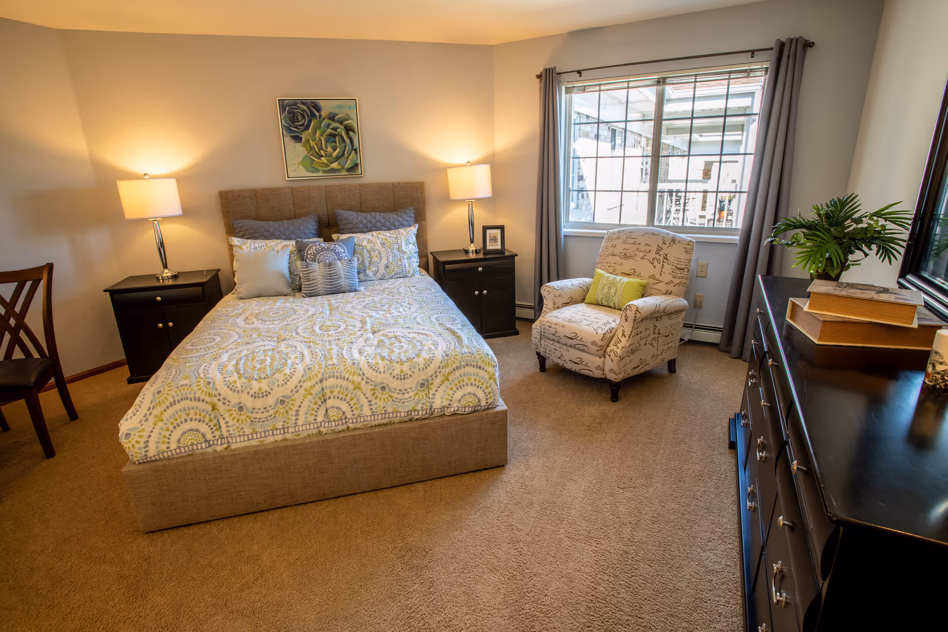 A cozy bedroom with a beige upholstered bed featuring patterned bedding and multiple pillows. There are two black nightstands with lamps on either side of the bed. A comfortable armchair with script print and a green pillow is placed near a window with gray curtains. A dark wooden dresser with books and a plant is on the right side of the room. The walls are light-colored and there is a framed succulent artwork above the bed.