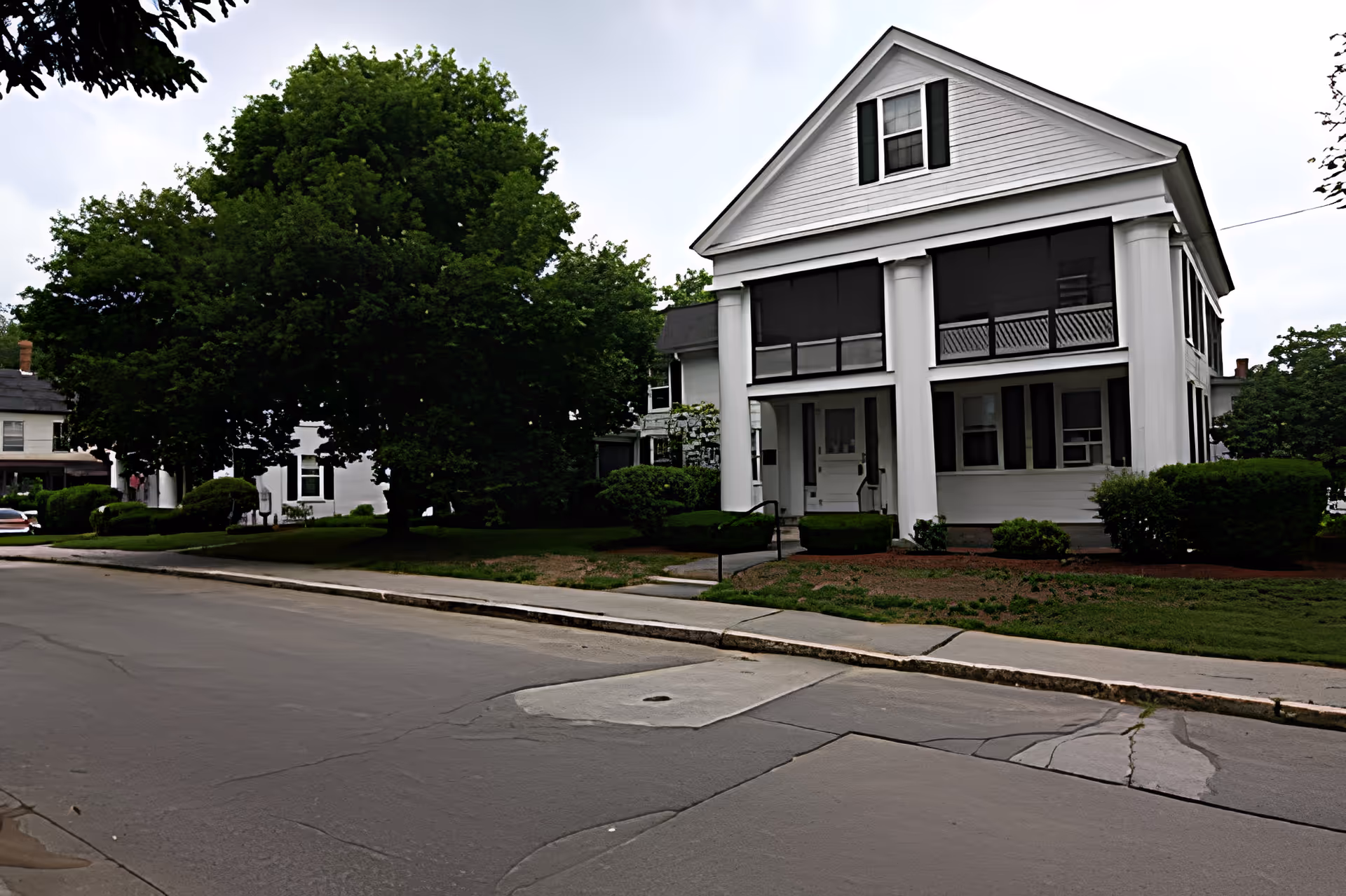 A large white two-story house with a gabled roof and prominent white columns at the front entrance. The house has a screened-in porch on the second floor and is surrounded by green bushes and trees. The street and sidewalk are visible in the foreground.