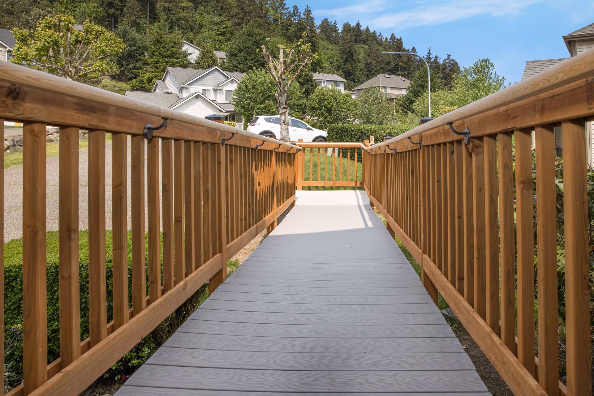 A wooden ramp with handrails on both sides leading to a small fenced area, surrounded by green grass and trees, with houses and a white car visible in the background under a clear blue sky.