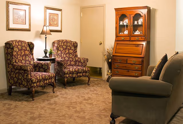 A cozy sitting area with two patterned armchairs, a small round table with a lamp and plant, a wooden cabinet displaying china, and a cushioned sofa with pillows, all set on a carpeted floor against beige walls.