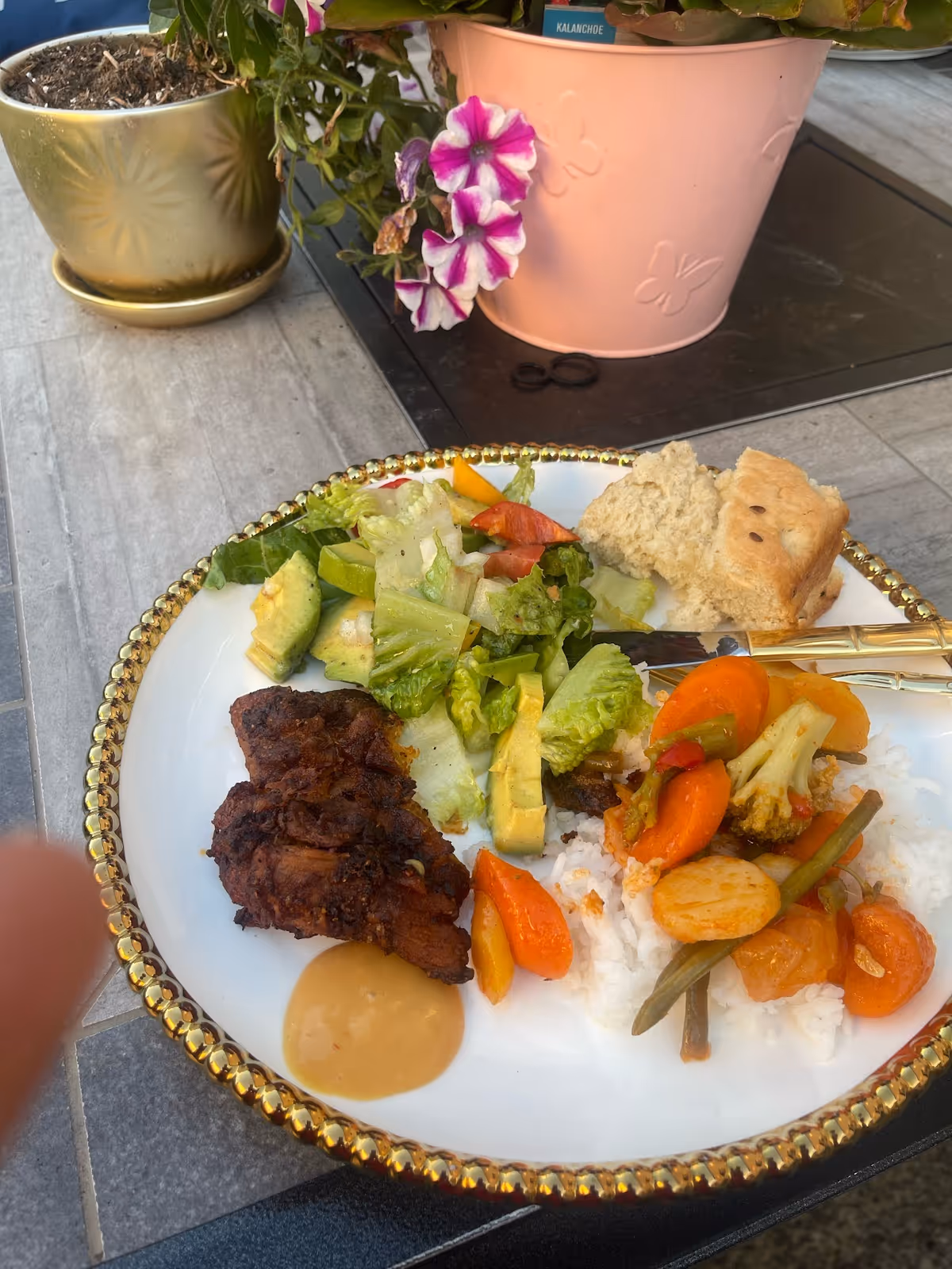 A plate of food on a table with a gold-rimmed edge, featuring grilled meat, a portion of salad with avocado, cooked mixed vegetables over white rice, a dollop of sauce, and two pieces of bread. In the background, there are two flower pots, one gold and one pink with purple and white flowers.