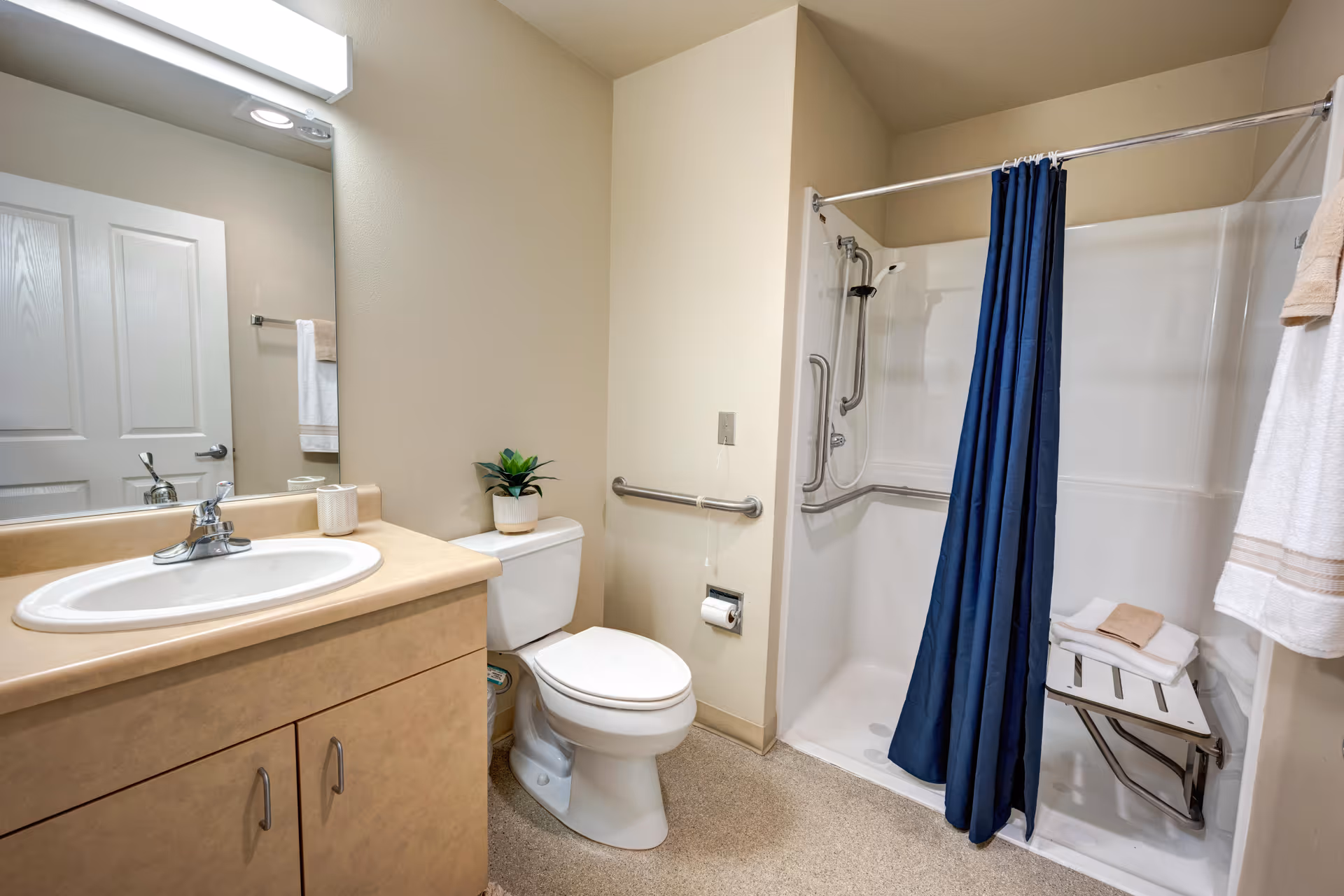 A clean and accessible bathroom featuring a beige countertop with a white sink, a toilet with a small potted plant on top, and a walk-in shower with grab bars, a fold-down seat, and a dark blue shower curtain. White towels hang on a rack beside the shower.