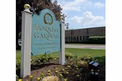A teal Hornell Gardens sign stands in a flower garden beside a brick building.