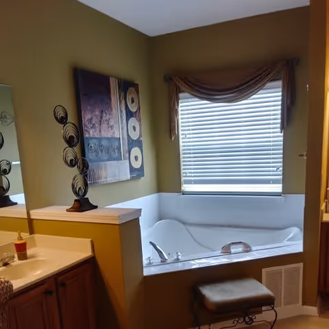 A bathroom with a large white bathtub under a window with closed blinds and a brown valance. To the left is a sink with a countertop and wooden cabinets below. Above the countertop is a decorative metal sculpture and a framed abstract painting on the wall. The walls are painted green with a white trim.