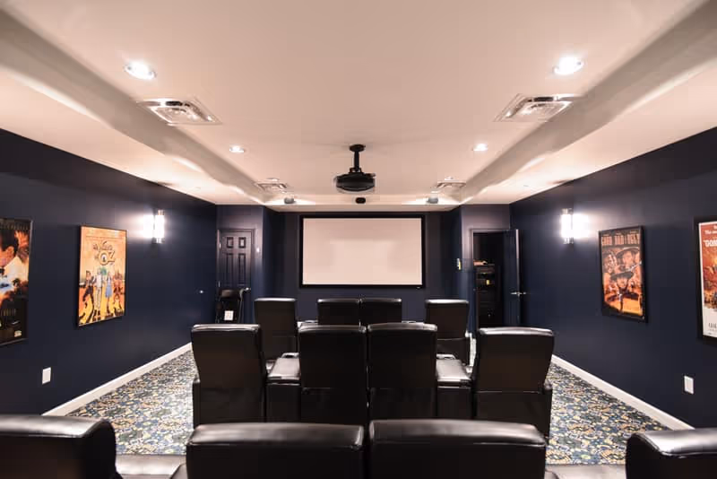 A cozy home theater room with multiple black leather recliner chairs arranged in rows facing a large white projection screen. The walls are painted dark blue and decorated with framed movie posters. The ceiling has recessed lighting and a mounted projector.