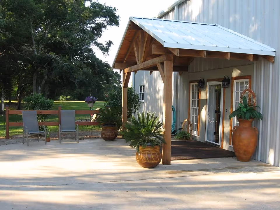 Outdoor patio area with a wooden pergola attached to a building. There are large potted plants and two chairs on a concrete surface. In the background, there is a wooden fence and green trees.