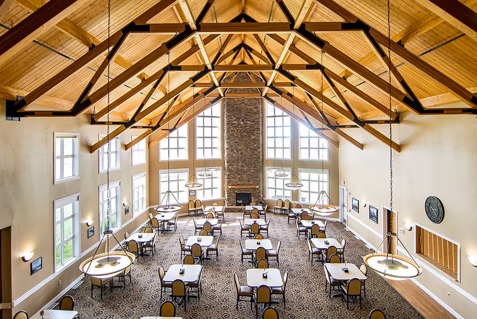 Spacious dining room with high vaulted wooden ceiling and exposed beams, multiple square tables each surrounded by four chairs, large windows letting in natural light, a stone fireplace at the far end, and hanging pendant lights.