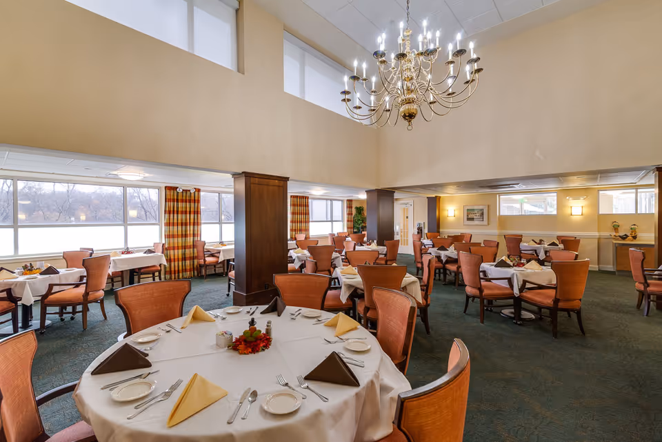 A spacious dining room with multiple round and rectangular tables covered with white tablecloths, each set with plates, silverware, and folded napkins in brown and yellow. The room features large windows with plaid curtains, green carpet, wooden columns, and a large ornate chandelier hanging from a high ceiling.