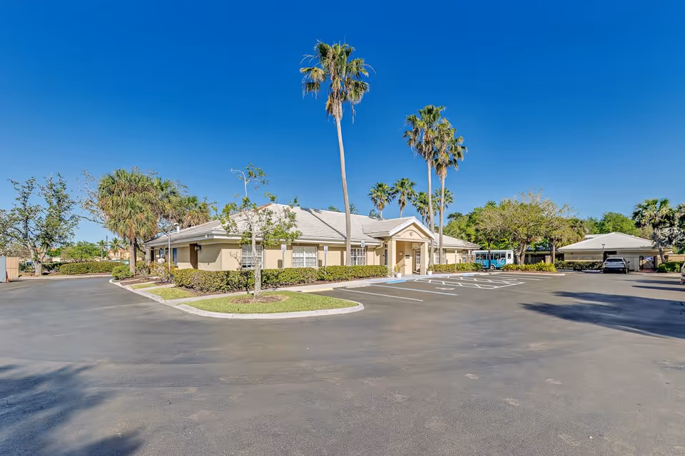 Exterior view of a single-story senior living facility building with a beige facade and a light-colored roof, surrounded by palm trees and other greenery under a clear blue sky. The building is adjacent to a large, mostly empty parking lot with marked parking spaces.