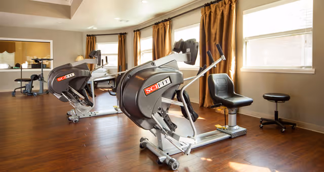 A well-lit exercise room with two SCI FIT stationary exercise machines positioned on a wooden floor. The room has large windows with brown curtains allowing natural light to enter. There is a small black stool near the window and a desk with a chair in the background.