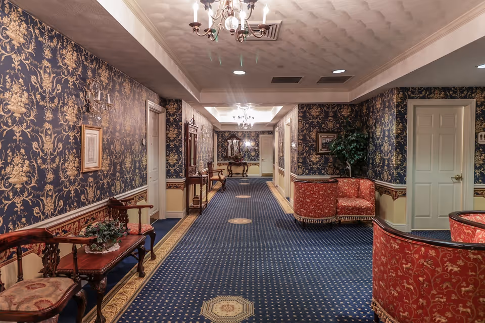 Carpeted interior hallway with ornate wallpaper, chandeliers, wooden benches and upholstered seating in a senior residence.