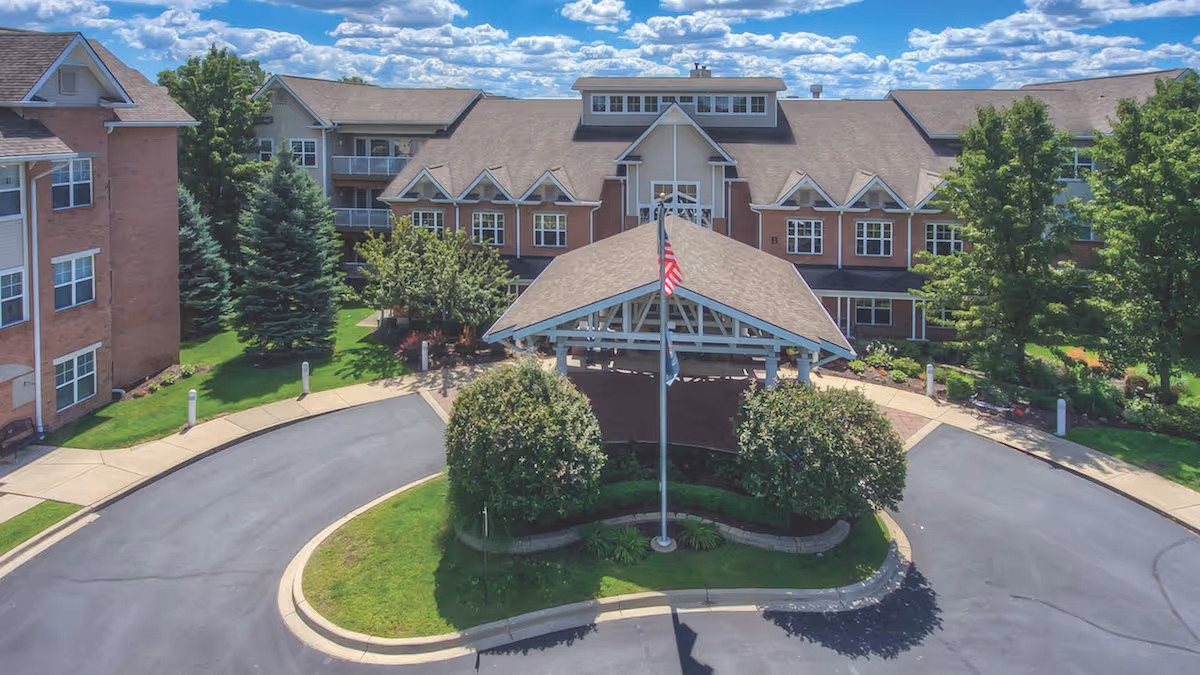 Front exterior view of Town Village Sterling Heights senior living facility with a covered entrance, American flag on a flagpole, well-maintained landscaping including trees and bushes, and a clear blue sky with scattered clouds.