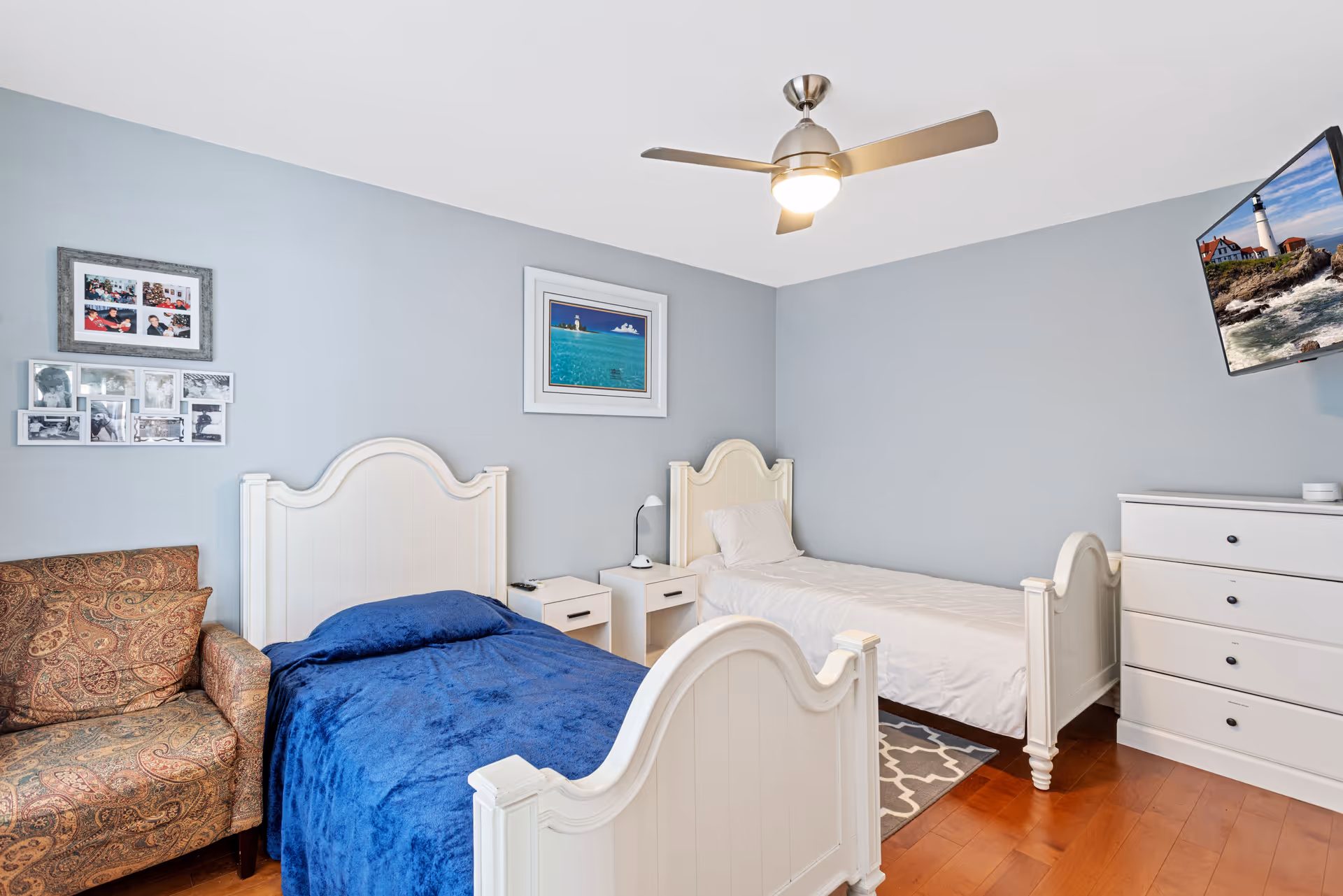 A bedroom with two white twin beds, one with a blue blanket and the other with a white blanket. Between the beds are two white nightstands with a small lamp on one. There is a patterned armchair on the left side and a white dresser on the right side with a wall-mounted TV above it displaying a lighthouse scene. The walls are painted light blue and decorated with framed pictures. A ceiling fan with a light is mounted on the white ceiling.