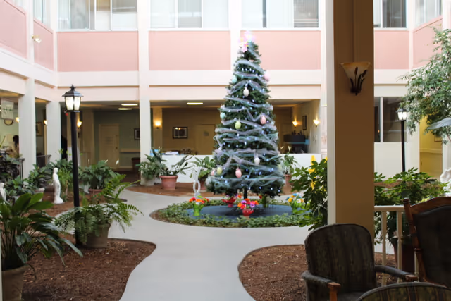Indoor courtyard area with a decorated Christmas tree in the center surrounded by plants and a circular walkway. There are chairs and potted plants around the space, with windows and doors leading to other rooms in the background.
