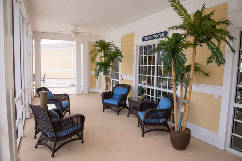 Covered porch with wicker chairs upholstered in blue, potted palm plants, and glass doors under a 'Bridgewater' sign.