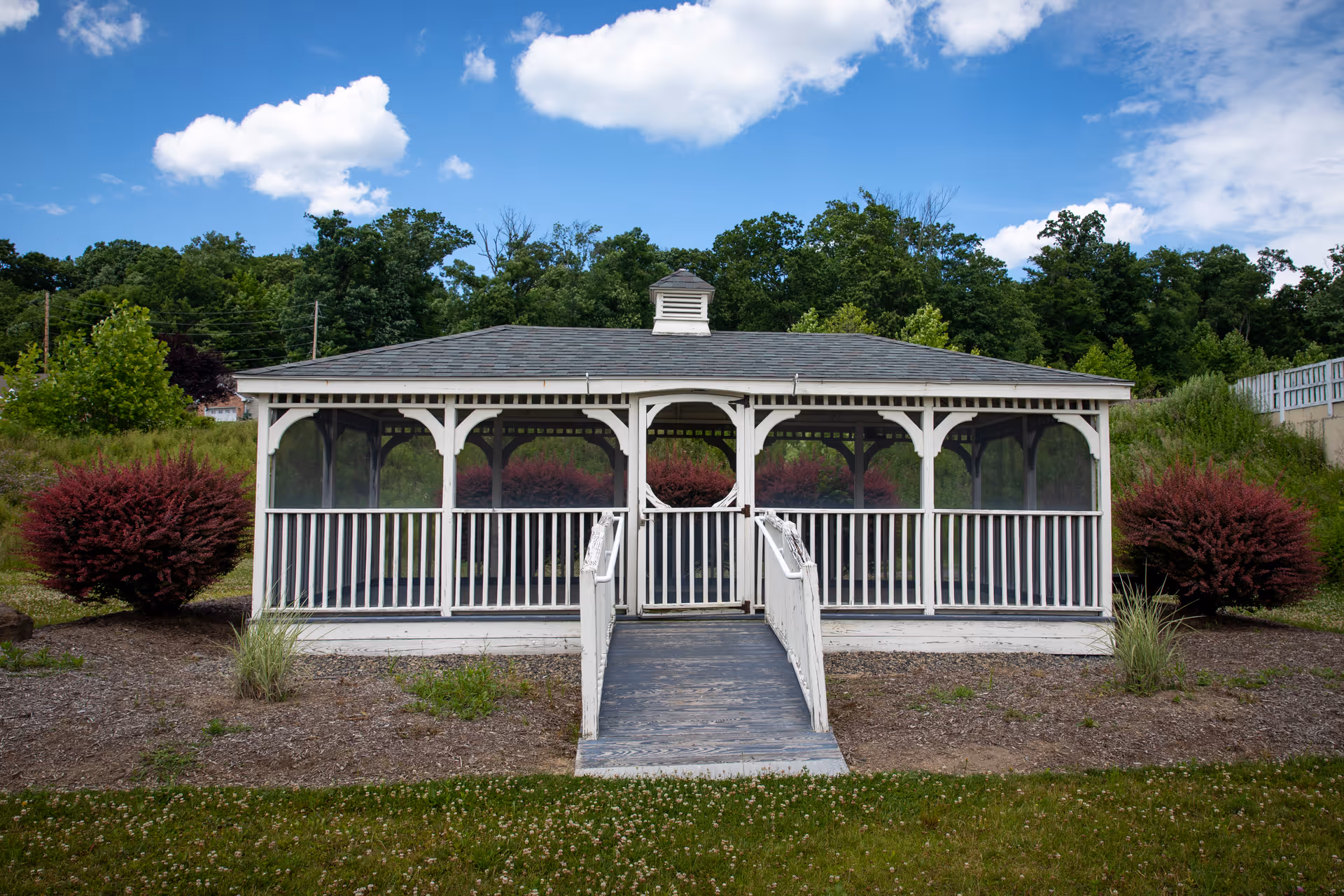 White wooden screened gazebo with a ramp on landscaped grounds under a blue sky.