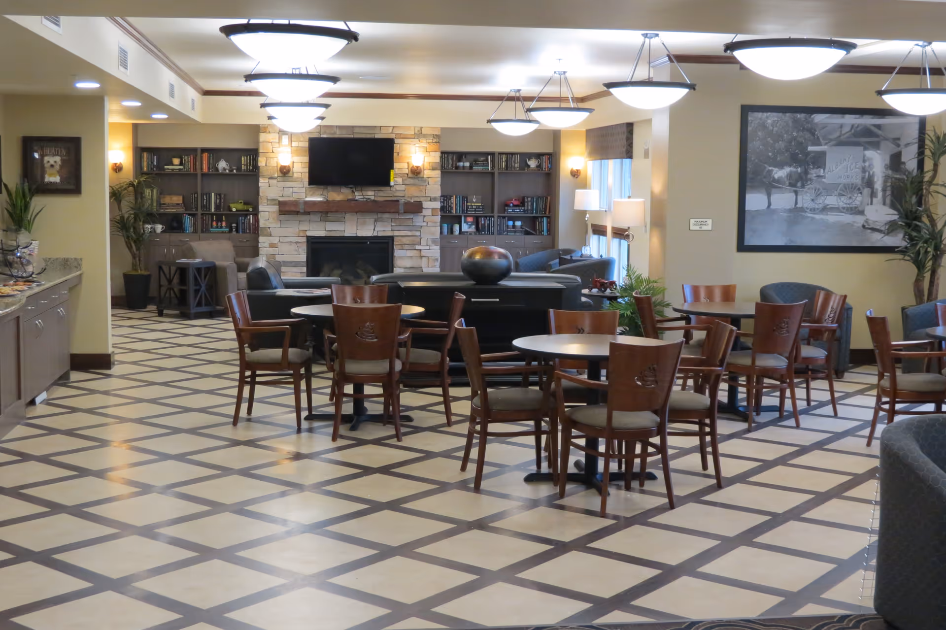 A spacious common area in a senior living facility featuring multiple round tables with wooden chairs, a stone fireplace with a mounted flat-screen TV, built-in bookshelves, and comfortable seating areas with armchairs and sofas. The room is well-lit with ceiling lights and wall sconces, and decorated with plants and framed artwork.