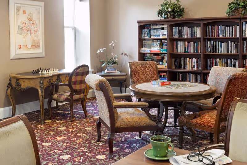 A cozy common area with a round table surrounded by four upholstered chairs, a bookshelf filled with books and board games, a small table with a chess set, and a window letting in natural light. The room has a patterned carpet and decorative plants on top of the bookshelf.