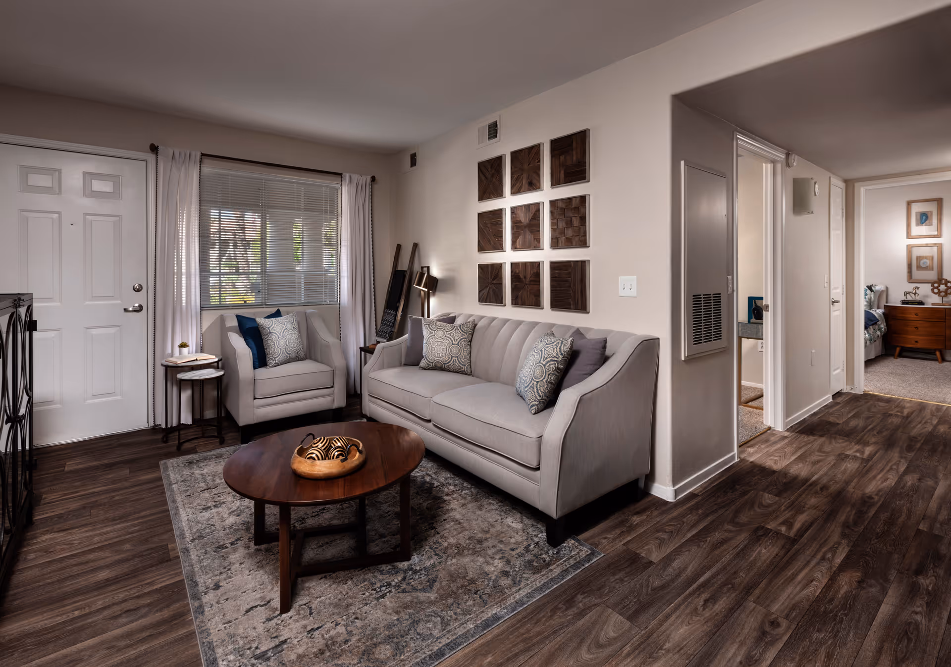 A cozy living room with a gray sofa and matching armchair, decorated with patterned pillows. A wooden coffee table with decorative items sits on a patterned rug. The room has wood flooring, a window with white curtains, and nine square wooden wall art pieces arranged in a grid. A hallway leads to a bedroom and bathroom.
