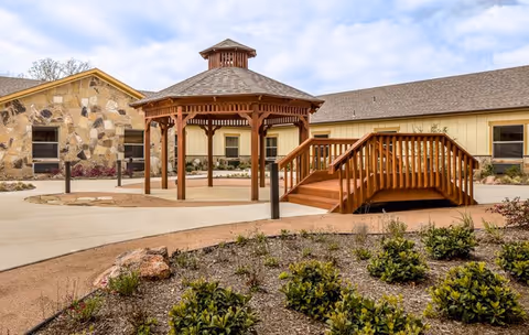 Outdoor courtyard area with a wooden gazebo and a small wooden bridge over a dry garden bed, surrounded by a beige building with stone and siding exterior walls under a partly cloudy sky.