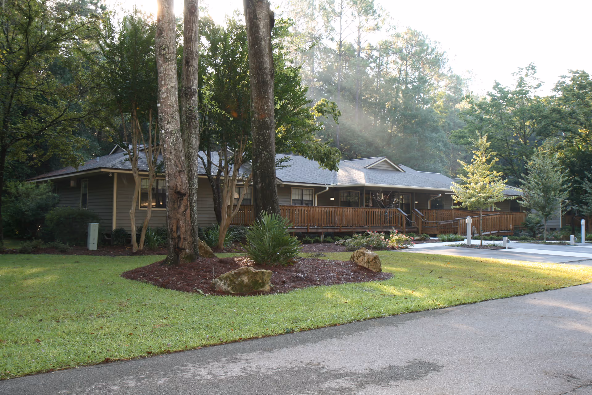 A single-story building surrounded by trees and greenery with a wooden ramp leading to the entrance, situated in a well-maintained outdoor area with grass, shrubs, and a paved driveway.