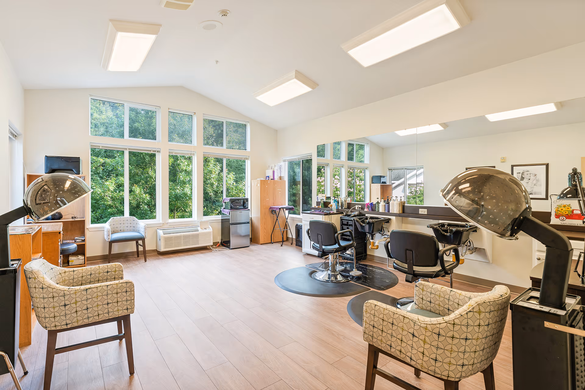 Bright salon-style interior with styling chairs, hooded hair dryers, wash basins, mirrors and lounge seating in front of large windows.