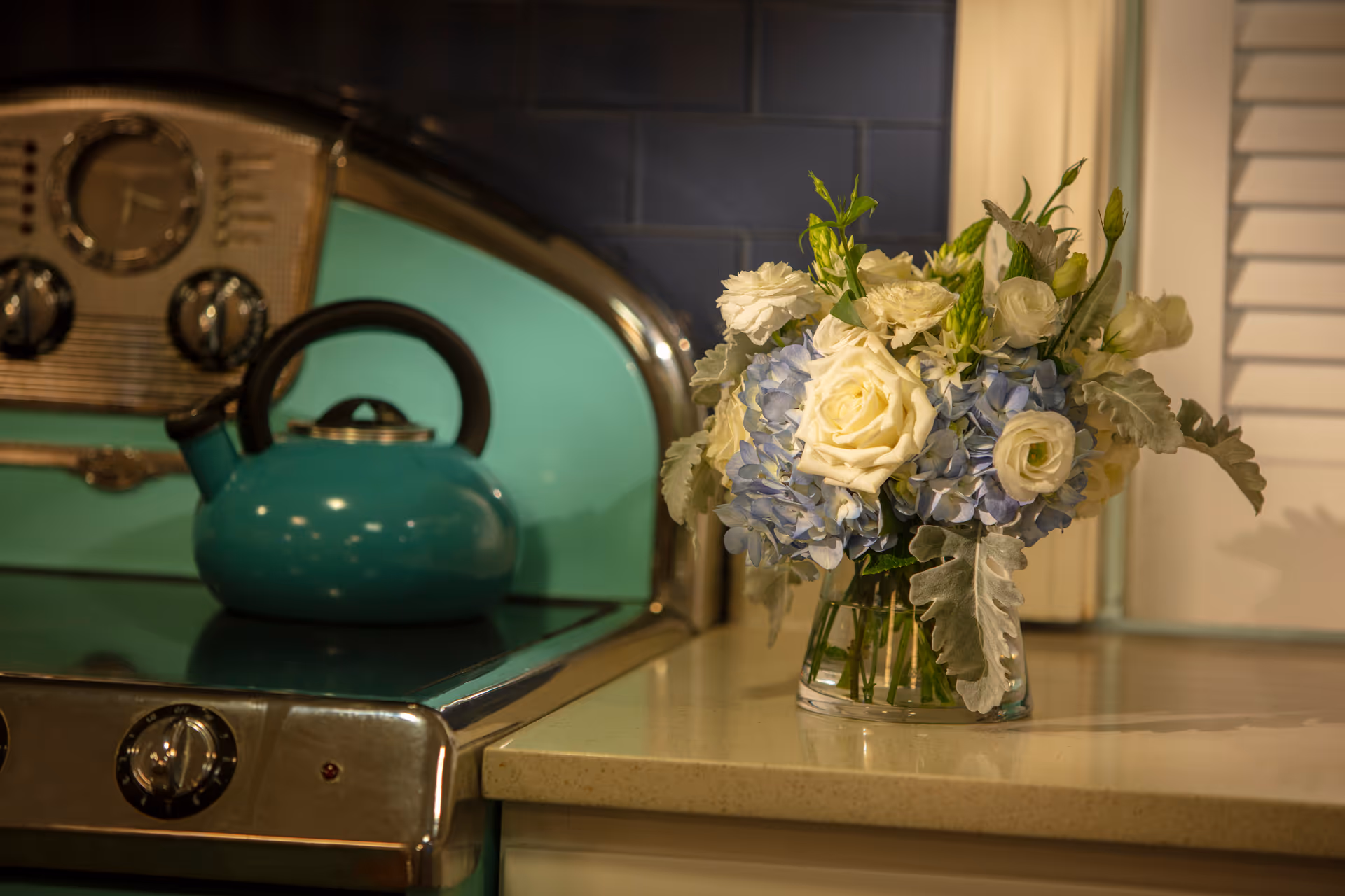 A vase of white and blue flowers sits on a kitchen counter beside a teal kettle on a retro stove.