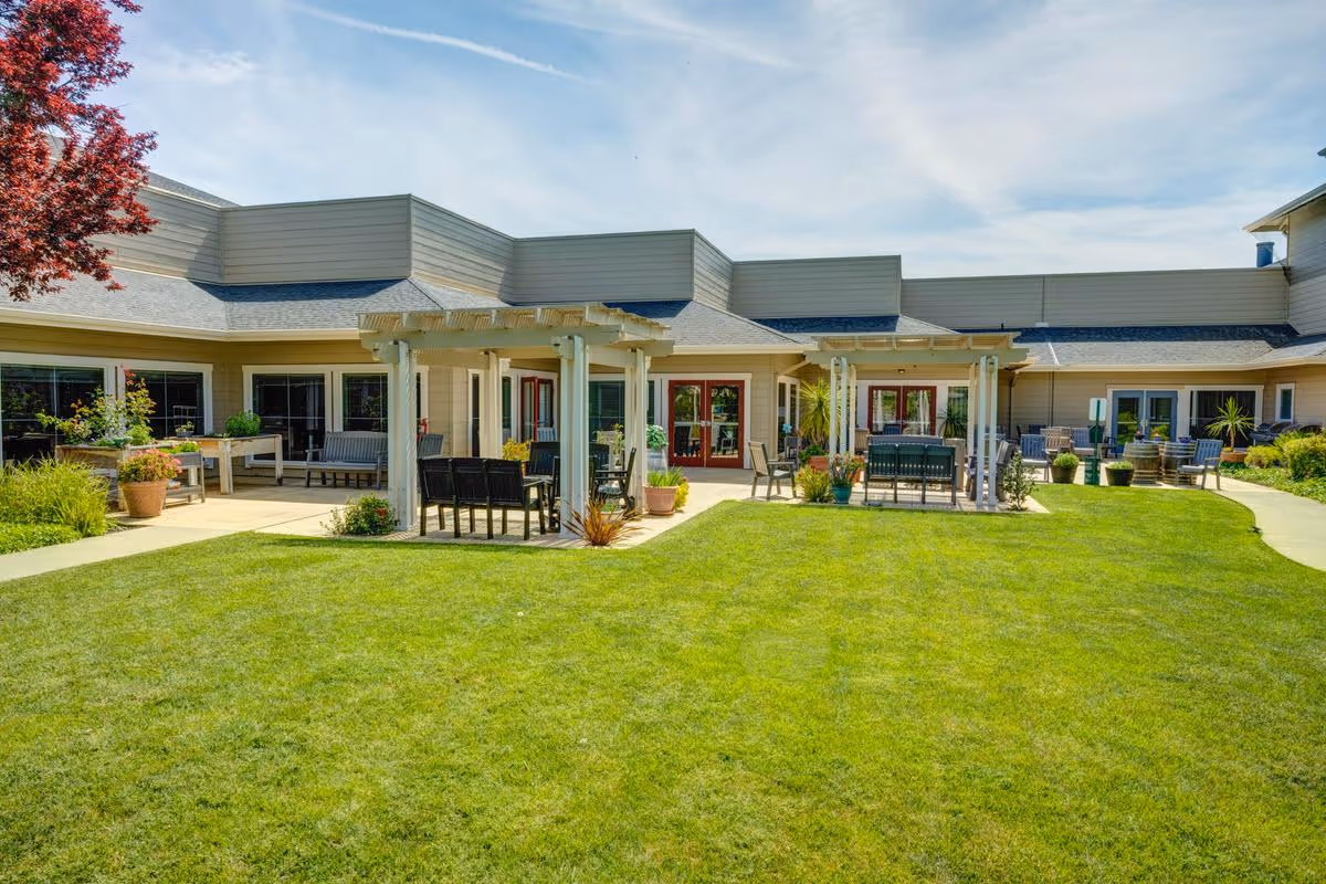 Outdoor courtyard area of a senior living facility with green grass, two white pergolas with seating underneath, potted plants, and a building with multiple windows and doors in the background under a partly cloudy sky.
