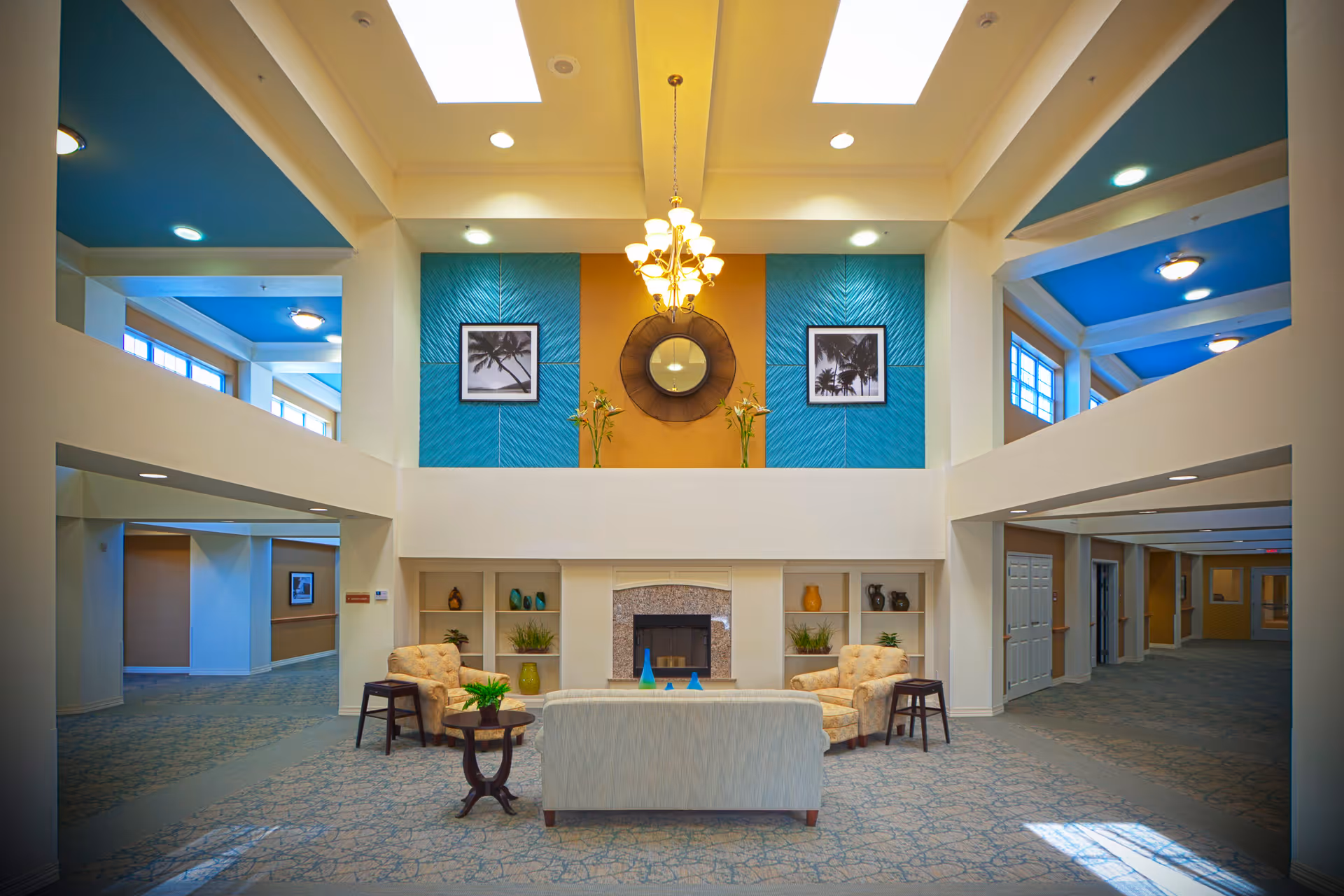 A spacious and well-lit common area in an assisted living facility featuring a fireplace with built-in shelves on either side, decorated with plants and vases. There are two beige armchairs and a light-colored sofa arranged around a small round table with a plant on it. The walls have blue and orange accent panels with framed black and white palm tree pictures and a round mirror. The ceiling has skylights and a chandelier, with open hallways visible on both sides.