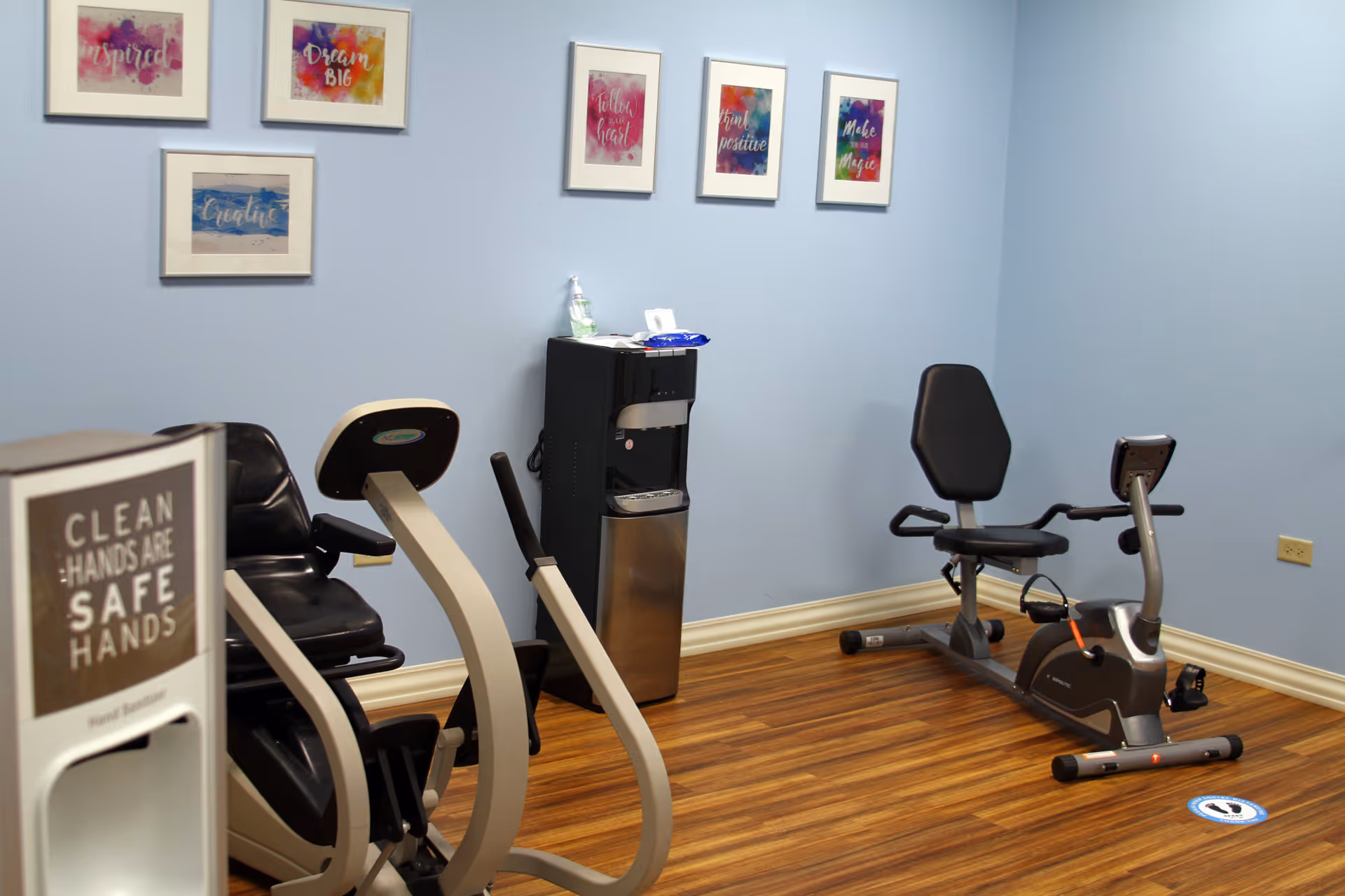 A small exercise room with two stationary recumbent bikes on a wood floor. The walls are light blue and decorated with six colorful framed motivational prints. There is a water dispenser with hand sanitizer and wipes on top. A sign in the foreground reads 'Clean Hands Are Safe Hands'.