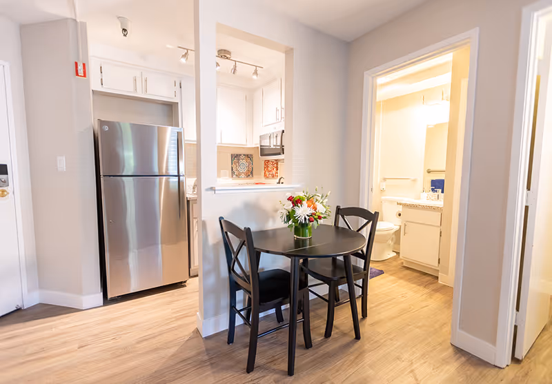 A small dining area with a round black table and two black chairs, a vase with flowers on the table. To the left is a kitchen with a stainless steel refrigerator and white cabinets. To the right is a bathroom with a toilet, sink, and mirror visible. The floor is light wood and the walls are painted white.