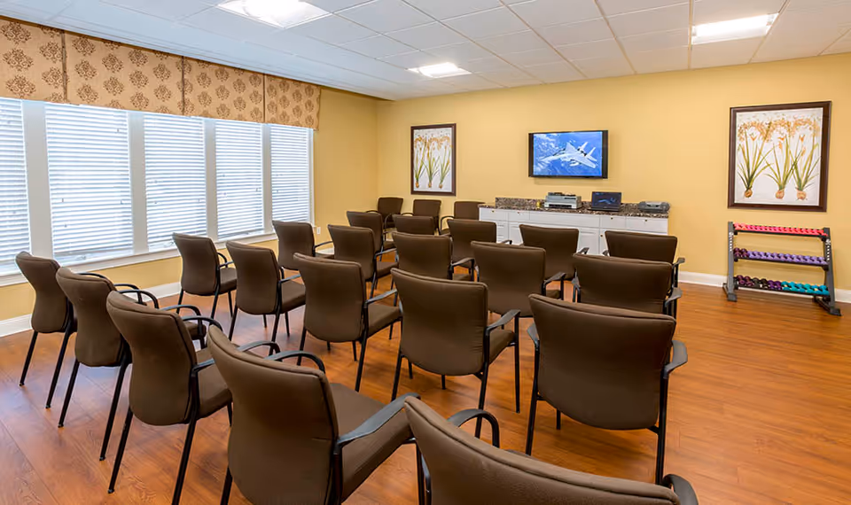 A room with multiple brown chairs arranged in rows facing a wall-mounted television. The room has wooden flooring, yellow walls, two framed botanical prints, a cabinet with electronic equipment, and a rack of colorful dumbbells in the corner. Large windows with blinds and a patterned valance allow natural light into the space.