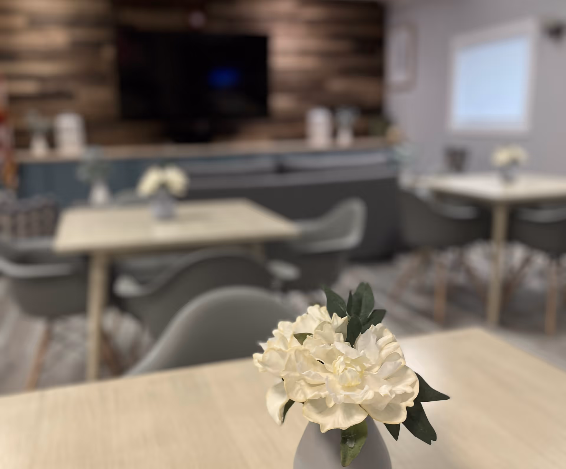 Interior of a dining area with light-colored tables and gray chairs. Each table has a small vase with white flowers. In the background, there is a wall-mounted TV and a wooden accent wall.